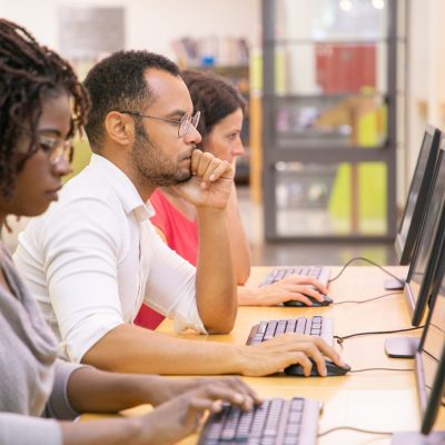 Multiracial group of students training in computer class. Line of man and women in casual sitting at table, using desktops, typing, looking at monitor. Training center concept