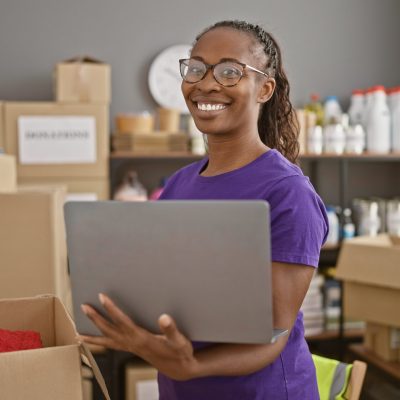 Smiling woman holding laptop in warehouse with donation boxes, portraying volunteer work and community service.